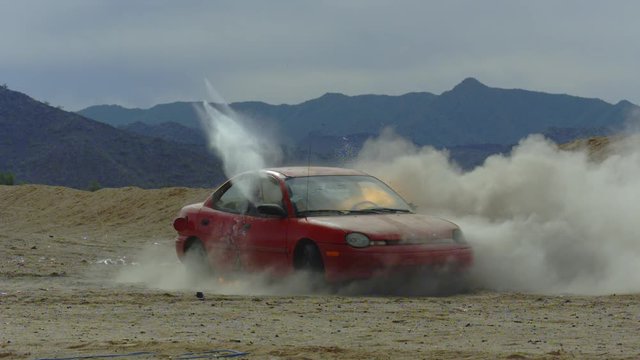 A wide shot of a red car shot up by bullets in slow motion.