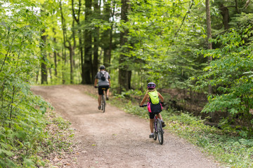 Couple of people cycling together in the woods down a dirt path on a beautiful summer day. Green canopy of trees above them. View from the back of people - full body.