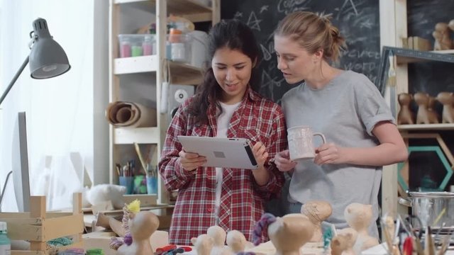 Young women standing in creative workshop filled with papier-mache figures chatting and looking at new project on tablet