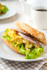 Sandwich stuffed with scrambled eggs, ham and lettuce leaves on a light wooden background. Selective focus.