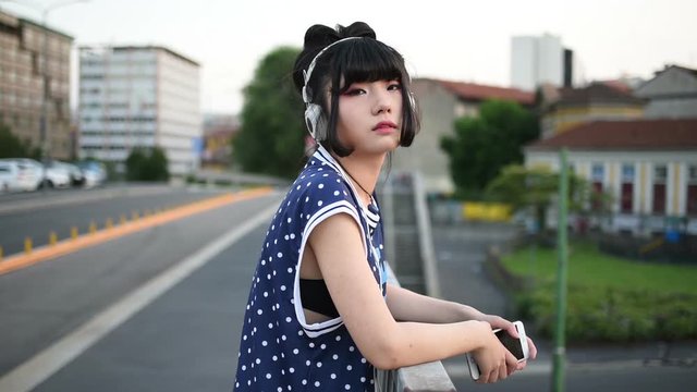 young beautiful asian millennial woman listening music with earphones and smart phone hand hold outdoor in city back light, looking at camera - relaxing, music, technology concept