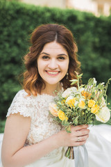 Beautiful brunette bride in elegant white dress holding bouquet posing neat trees