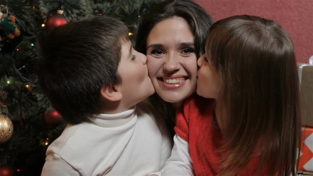 Caucasian children kissing their mother from both sides. Pretty young woman sitting between little boy and girl. Attractive brunette woman with young kids smiling for the camera