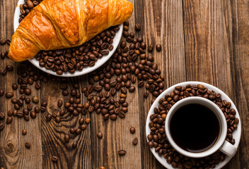 Breakfast coffee - cup and croissant with coffee beans on wooden background.