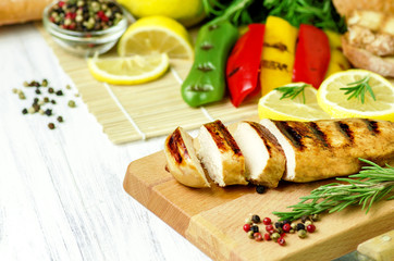 Grilled chicken breast placed on wooden board with grilled sweet colored pepper ,mixed peppers, rosemary,bread and lemon on white wooden background.