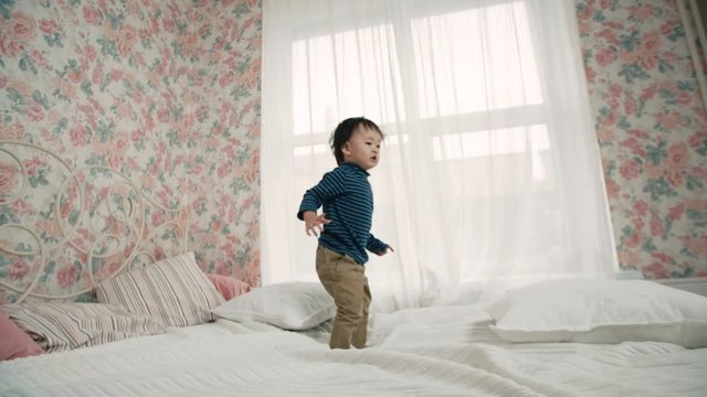 the little boy of Asian appearance, having fun on the bed in the room, laughing and jumping, slow motion