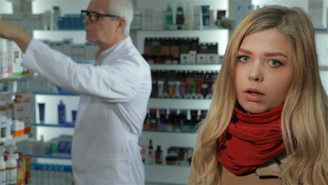 Pretty blond woman caughing at the drugstore. Young caucasian woman in red big scarf needs remedy for caugh at the pharmacy. Attractive female customer standing against background of male pharmacist