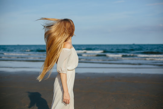 Enjoyment, free happy woman enjoying sea beach. Beautiful blond woman in white dress walking on sunset, enjoying peace, serenity in nature