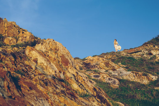 Elegant young woman in white dress on top of mountain cliff. Enjoying the freedom of travel, nature, blue sky