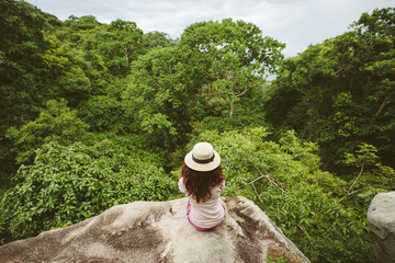 Young woman in white hat sitting back on tropical mountain cliff