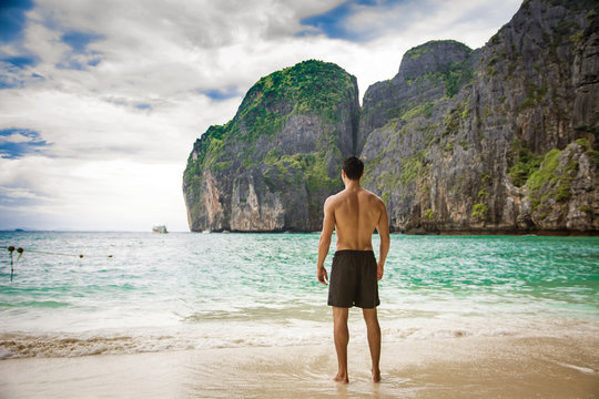 Full body shot from the back of a handsome young man standing on a beach in Phuket Island, Thailand, shirtless wearing boxer shorts, showing muscular fit body