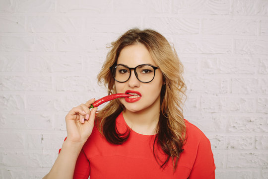 Beautiful casual caucasian woman wearing red dress and glasses with chili pepper in mouth