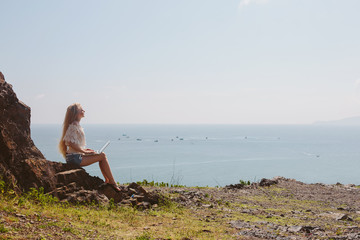 Young attractive woman sitting outdoor near beautiful ocean using laptop computer and enjoy freedom and summer vacation