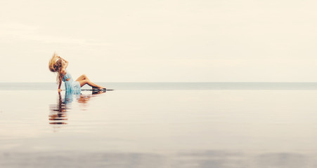 Fototapeta premium Young female sitting on the edge of swimming pool. Beautiful sea water skyline. Spa treatments, nature, freshness of water and relaxation