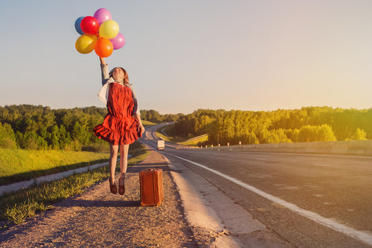 Young happy woman jumping with coloured balloons. Large suitcase, freedom of travel, road