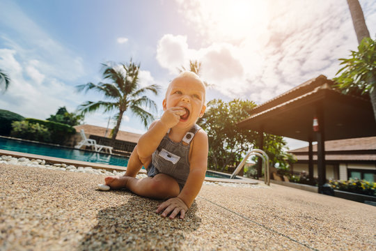 Funny baby boy trying to  eat stone outdoor near swimming pool. Summer vacation, sunny day, palm trees