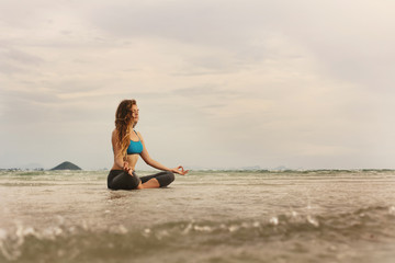 Fototapeta na wymiar Young caucasian Woman sitting on sea shore and practicing yoga meditation. Enjoy ocean waves, wind and sun