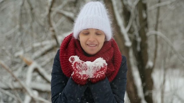 young girl in white knitted hat, red scarf and mittens blows snow with hands in snowy woods