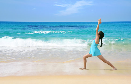 Young healthy Asian kid doing yoga on tropical beach in warrior pose I. Preteen girl in swimwear practice yoga on sea beach blue sky sand sun daylight relaxation landscape for sun protection ad.