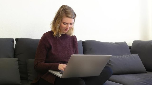 Junge europ&auml;ische Frau sitzt auf dem Sofa und schreibt mit einem Laptop und hat gute Laune