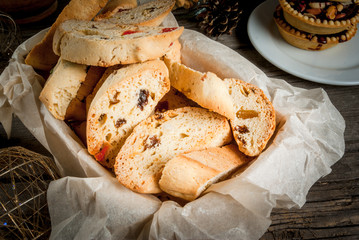 Traditional Christmas baking biscotti or cantucci in a basket on a wooden table next to the branches of a fir-tree, decorations and other kinds of Christmas sweets (tarts with nuts, ginger biscuits)