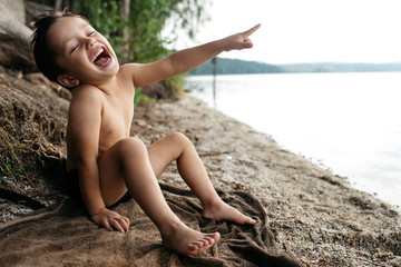 little boy is 3-4 years old sitting on the lake shore, dressed in swimming shorts. He laughs and points a finger, grimaces