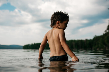 Little boy 4 years is waist-deep in the water of  forest lake. View from the back. Beautiful nature, water surface, forest and mountains on the horizon