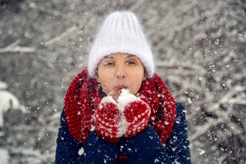 young girl in white knitted hat, red scarf and mittens blows snow
