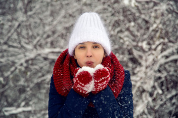 young girl in white knitted hat, red scarf and mittens blows snow