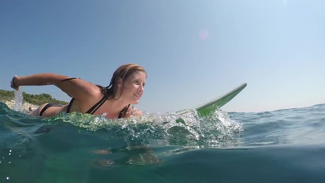 SLOW MOTION CLOSE UP, UNDERWATER: Happy female surfer lying on surfboard and paddling out on stunning sunny day. Excited girl on active summer vacation swimming and surfing in crystal clear blue ocean