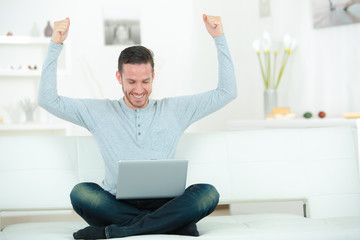 cheerful young man with laptop raising hands indoors