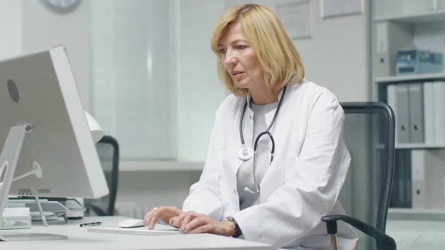 In the Doctor's Office. Mid Adult Female Doctor Works at Her Desktop Computer. In the Background Her Assistent's Works at His Desk.
 Shot on RED Cinema Camera in 4K (UHD).