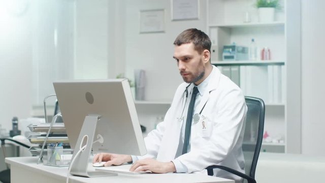 Male Doctor is Working at His Desk. He Uses Personal Computer and Consults Documents. Shot on RED Cinema Camera in 4K (UHD).
