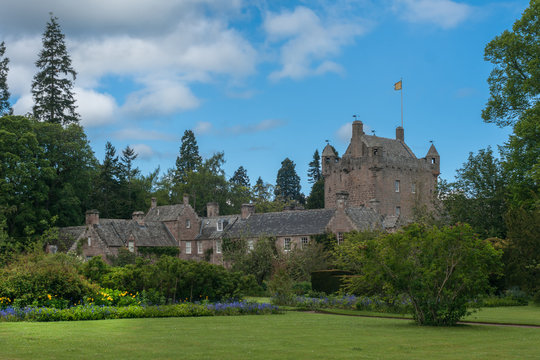 Cowdor, Scotland - June 2, 2012: The brown-gray stone structure of undamaged Cowdor Castle under blue sky. Flag on top. Large part of formal garden in forefront. Green foliage.