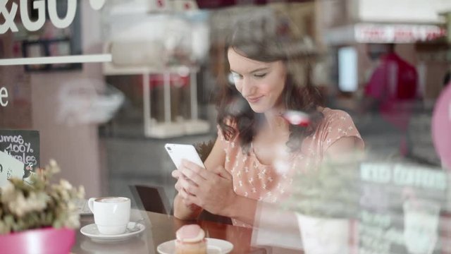 Young woman with her smartphone next to the window in a coffee shop. She smiles while texting. We see traffic and people in the reflection of the window.