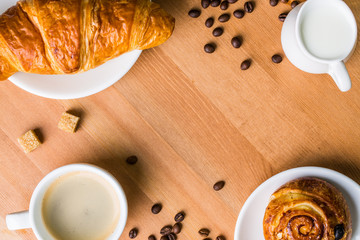 Coffee cup with milk jug, cinnabon and croissant on wooden background.