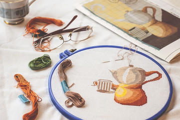 Cross-stitch set : hoop with embroidered teapot pattern, glasses, scheme, scissors, canvas and colorful yarn. Selective focus. Freelance, hobby, handmade home decor concept