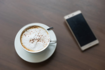 hot coffee in white cup white foam and mobile phone with silver spoon on wooden table at coffee time sunset / hot coffee mobile phone