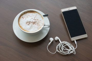 hot coffee in white cup white foam and mobile phone and headphones with silver spoon on wooden table at coffee time sunset / hot coffee mobile phone