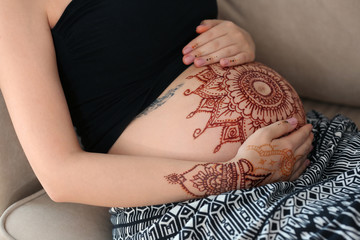 Young pregnant woman with henna tattoo on belly sitting on sofa in the room