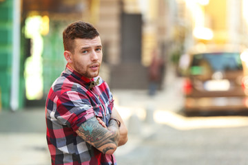Fototapeta premium Young man with tattooed arm and plaid shirt looking back on city street, he has a short beard and dark hair, the background is blurred with urban elements and sunlight