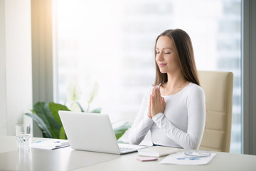 Young woman meditating sitting at the modern office desk in front of laptop, taking a pause, busy, stressful office, cure for work overload, one moment meditation, worshiping laptop