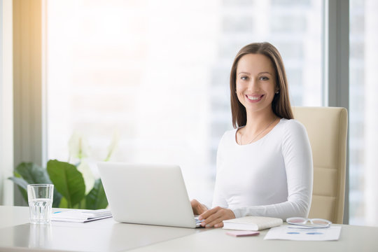 Young smiling female receptionist at the modern office desk with a laptop ready to greet clients, customers and visitors, direct them, free consultations, first impression. Looking at the camera