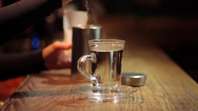 Footage of a woman placing a glass of hot water on a counter, in a bar, and adding a tea bag.
