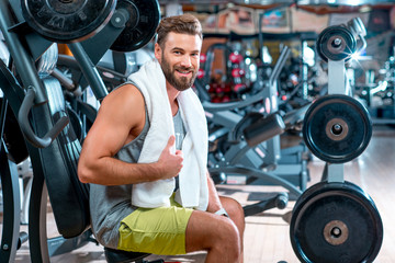 Fototapeta na wymiar Lifestyle portrait of handsome muscular man with towel sitting on the simulator in the gym