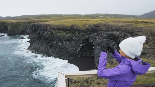 Tourist on travel taking photo with smart phone on Iceland of dramatic coast and ocean. Happy woman sightseeing taking pictures using smartphone visiting Arnarstapi, Snaefellsnes, West Iceland.