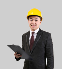 Handsome Asian man with clipboard and hardhat on light background