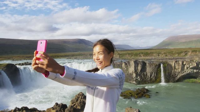 Tourist taking selfie photo with smart phone against majestic Godafoss waterfall. Happy young woman tourists enjoying icelandic nature landscape visiting famous tourist destination attraction, Iceland