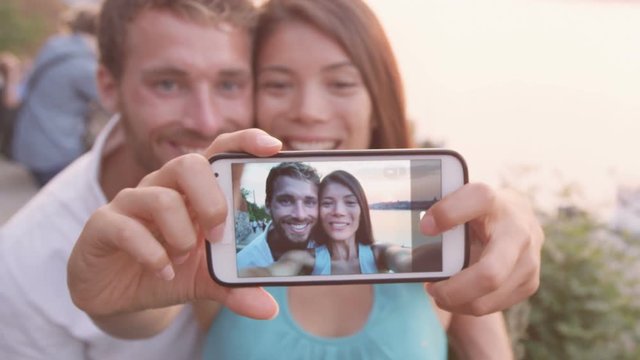 Smart phone selfie - couple taking self portrait using smartphone camera. Dating couple in love having fun taking candid fresh picture photo laughing smiling. Caucasian man  Asian woman at sunset.