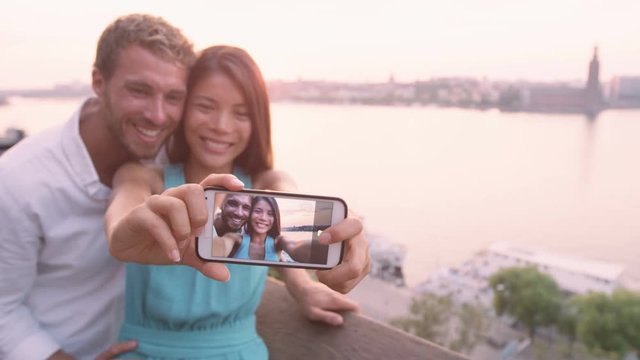 Selfie couple taking smart phone self portrait in Stockholm. People having fun with smartphone taking candid fresh selfportrait picture photo laughing smiling. Scandinavian man  Asian woman at sunset.